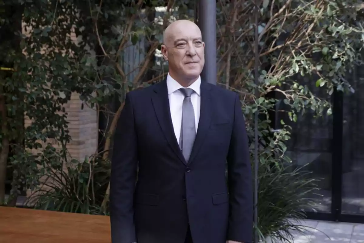 Hombre calvo con traje oscuro y corbata gris posando al aire libre frente a plantas y un edificio Hombre calvo con traje oscuro y corbata gris posando al aire libre frente a plantas y un edificio