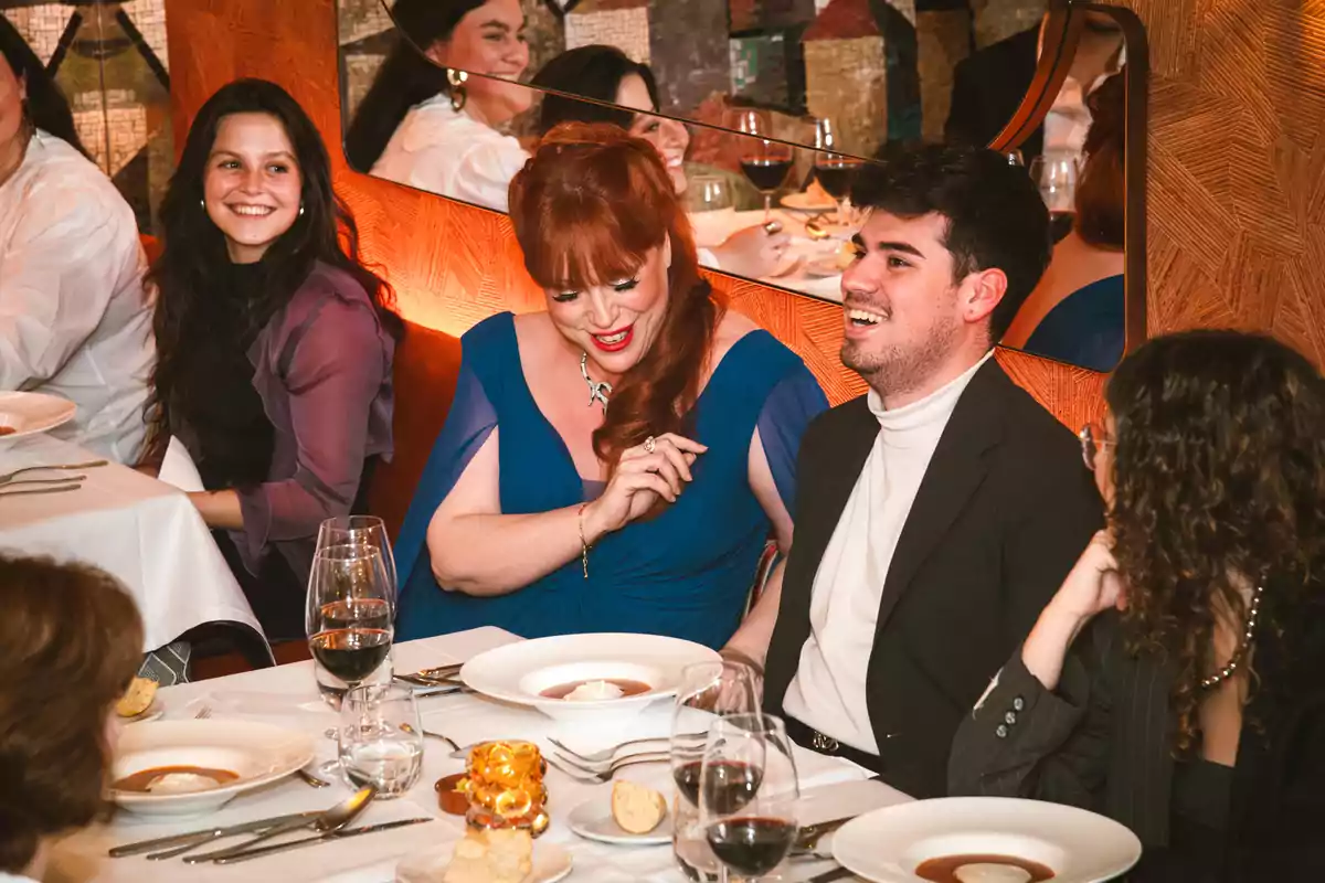 Varias personas elegantemente vestidas disfrutan de una cena en un restaurante, conversando y riendo alrededor de una mesa con platos y copas de vino. Varias personas elegantemente vestidas disfrutan de una cena en un restaurante, conversando y riendo alrededor de una mesa con platos y copas de vino.