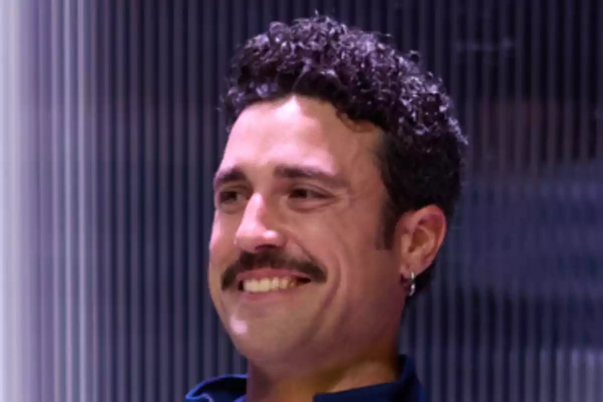 Hombre de cabello rizado y bigote sonriendo frente a un fondo de líneas verticales Hombre de cabello rizado y bigote sonriendo frente a un fondo de líneas verticales
