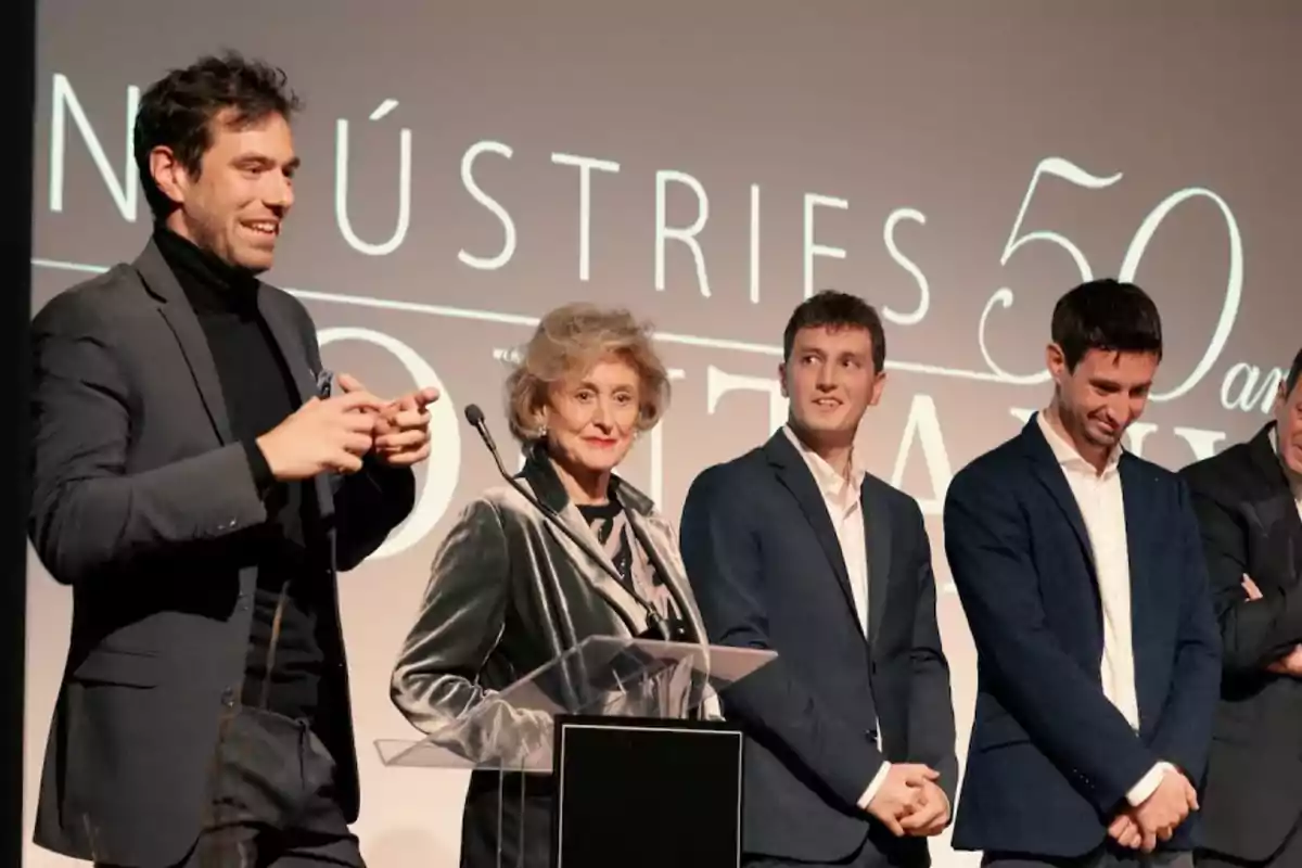 Cinco personas elegantemente vestidas están de pie en un escenario durante un evento, una mujer mayor está en el centro frente a un atril y los demás la acompañan sonrientes