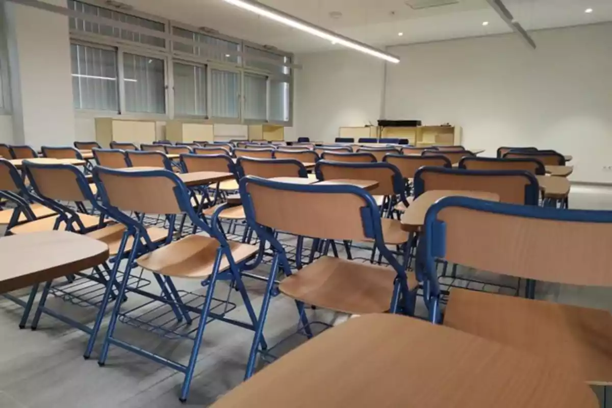 Aula vacía con varias filas de pupitres y sillas de madera con estructura azul frente a una pizarra y ventanas grandes