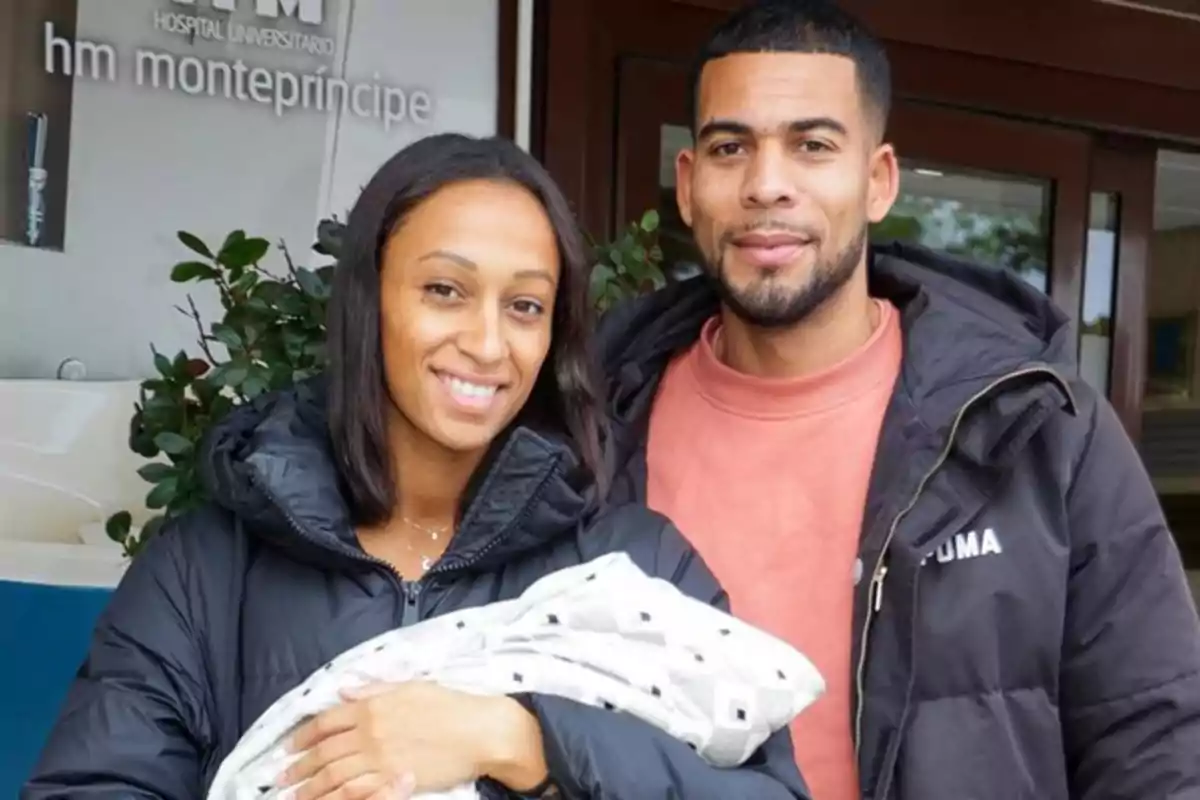 Una pareja sonriente sostiene a un bebé envuelto en una manta frente a la entrada de un hospital Una pareja sonriente sostiene a un bebé envuelto en una manta frente a la entrada de un hospital