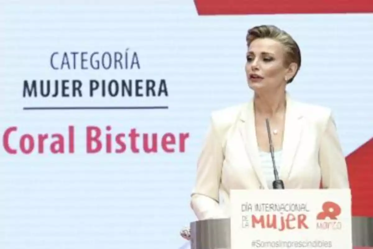 Mujer de cabello corto y rubio vestida con traje claro hablando en un podio durante un evento del Día Internacional de la Mujer donde se le reconoce en la categoría Mujer Pionera