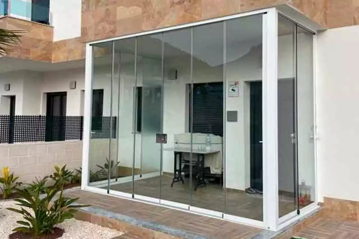 Terraza cerrada con paneles de vidrio junto a una casa moderna con plantas decorativas en el exterior