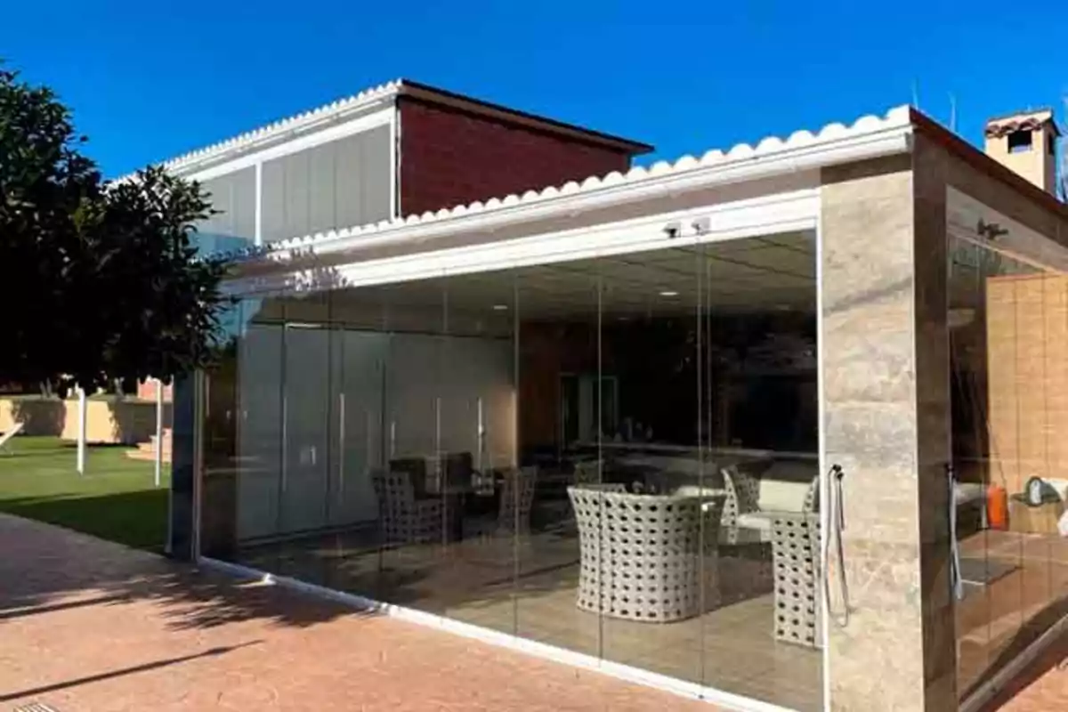Terraza cerrada con paredes de cristal y muebles de exterior en un jardín junto a una casa moderna Terraza cerrada con paredes de cristal y muebles de exterior en un jardín junto a una casa moderna