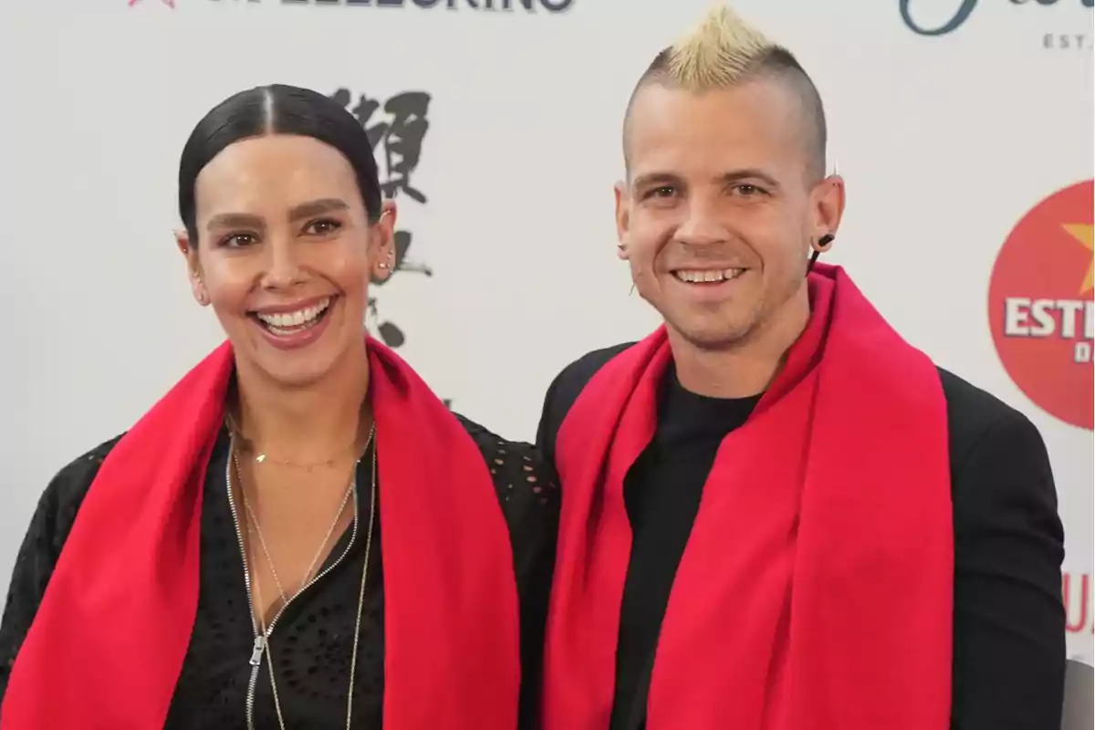 Dos personas sonrientes posan juntas en un evento, ambas llevan bufandas rojas sobre ropa oscura y están frente a un fondo con logotipos. Dos personas sonrientes posan juntas en un evento, ambas llevan bufandas rojas sobre ropa oscura y están frente a un fondo con logotipos.