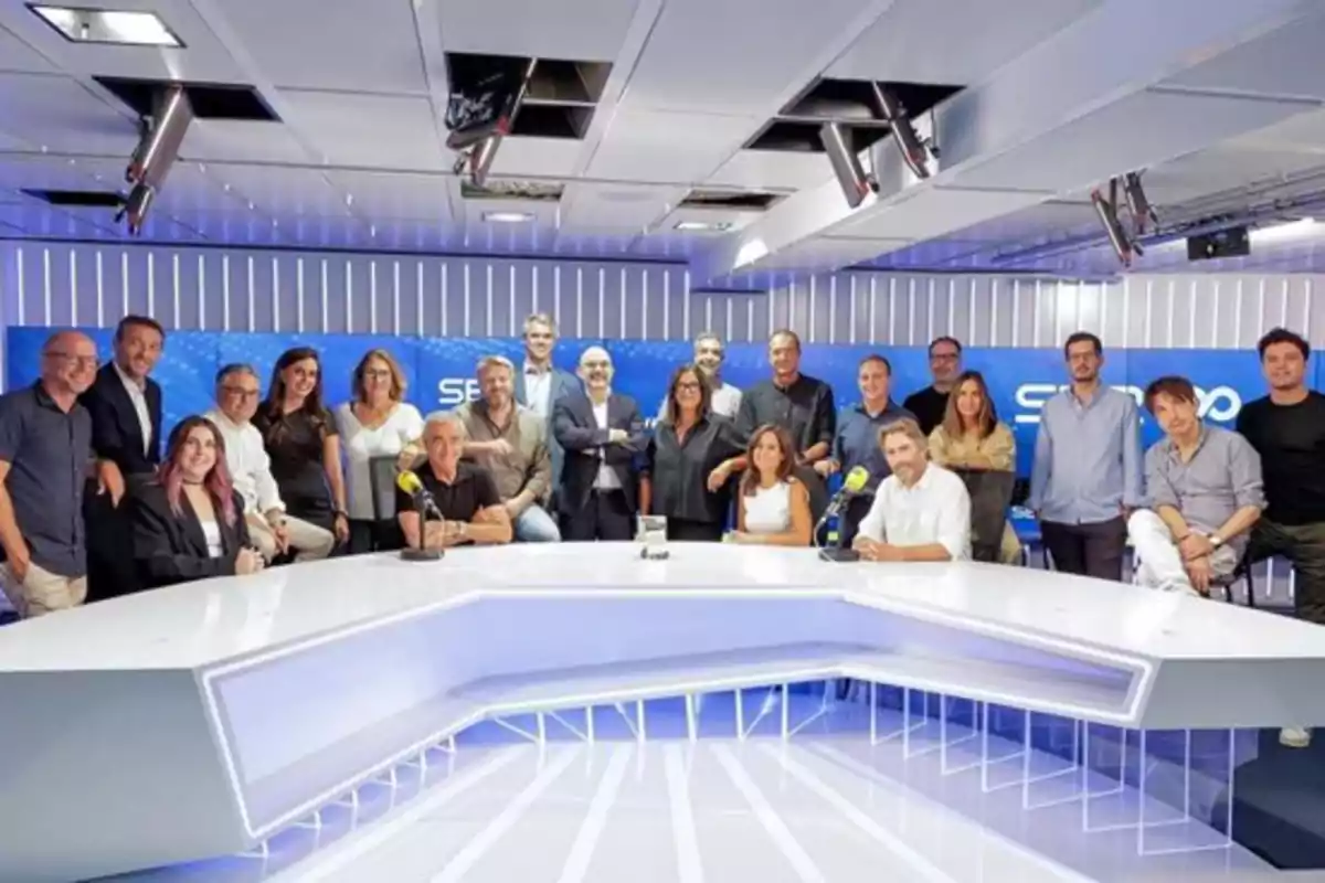 Un grupo numeroso de personas posando en un moderno estudio de radio con una mesa blanca y micrófonos amarillos Un grupo numeroso de personas posando en un moderno estudio de radio con una mesa blanca y micrófonos amarillos