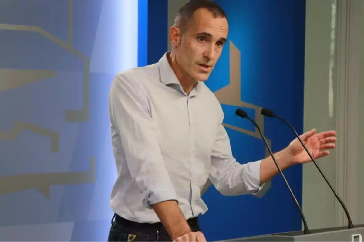 Hombre de pie hablando en una conferencia frente a dos micrófonos con fondo azul y gris Hombre de pie hablando en una conferencia frente a dos micrófonos con fondo azul y gris