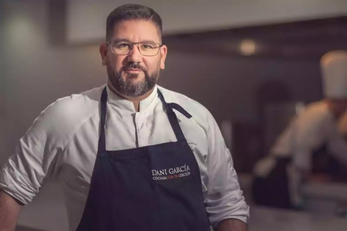 Chef con gafas y barba lleva un delantal oscuro y una chaqueta blanca de cocina mientras posa en una cocina profesional. Chef con gafas y barba lleva un delantal oscuro y una chaqueta blanca de cocina mientras posa en una cocina profesional.