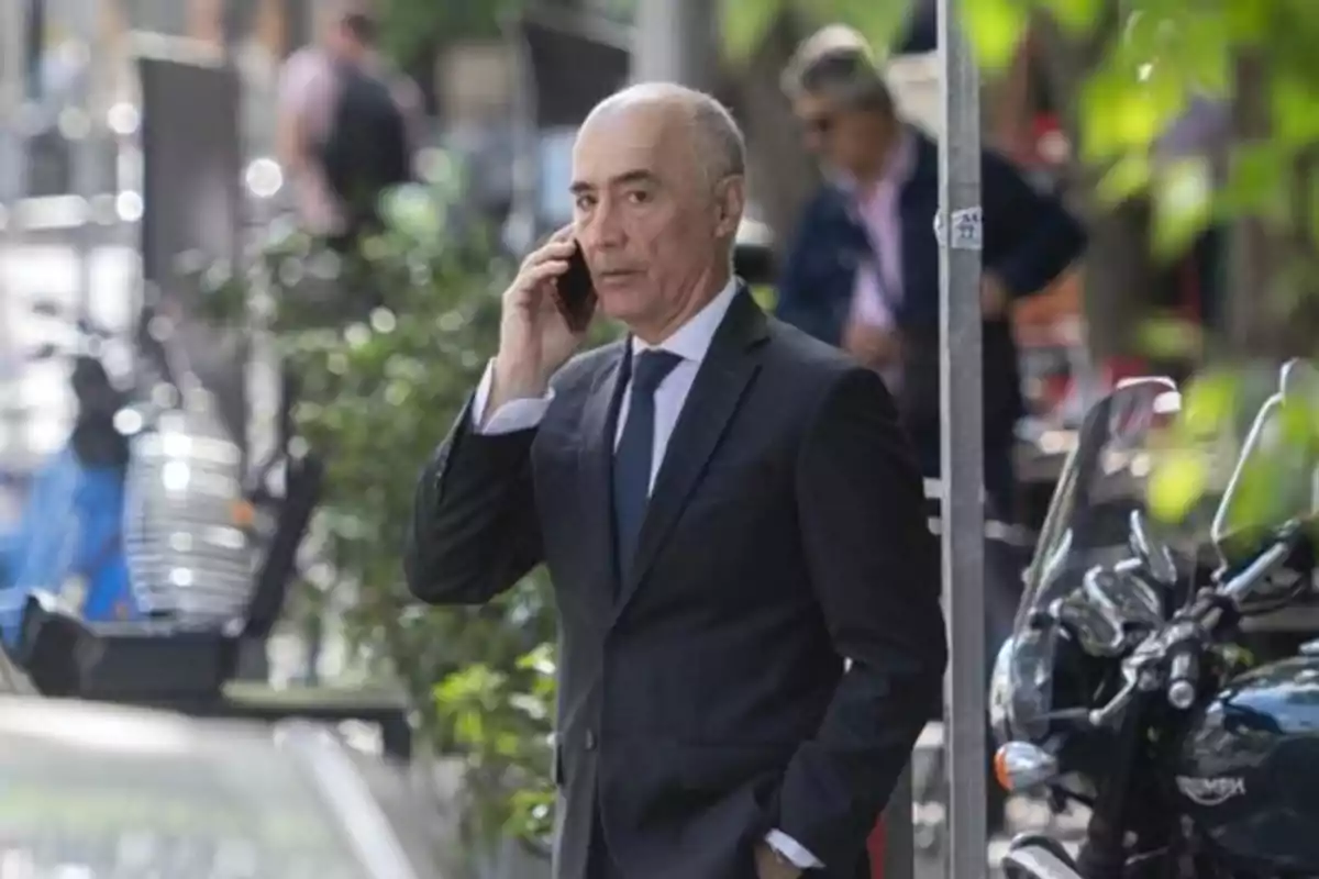Hombre de traje hablando por teléfono móvil en una calle urbana con motocicletas y vegetación alrededor Hombre de traje hablando por teléfono móvil en una calle urbana con motocicletas y vegetación alrededor