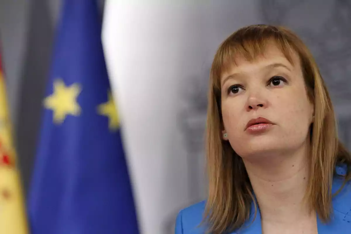 Mujer de cabello rubio y liso con chaqueta azul frente a banderas de España y la Unión Europea Mujer de cabello rubio y liso con chaqueta azul frente a banderas de España y la Unión Europea