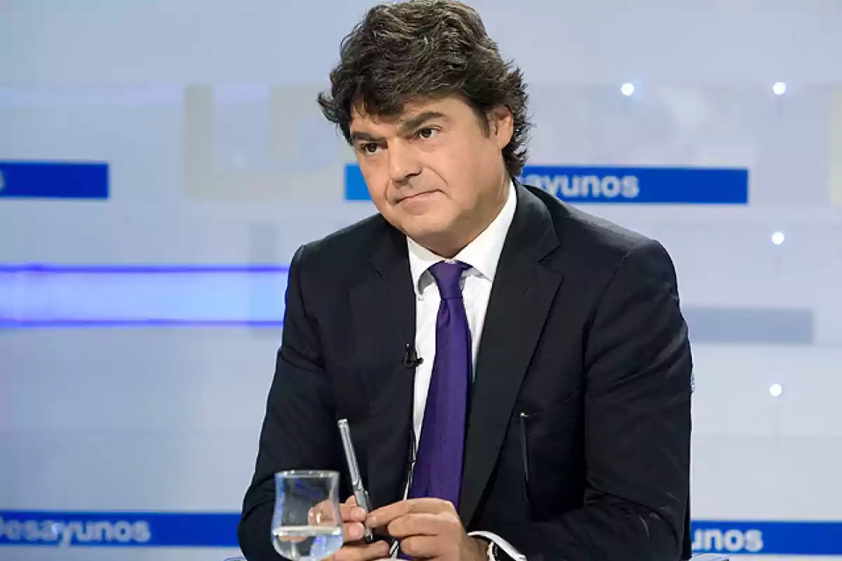 Hombre de cabello oscuro con traje y corbata morada sentado en un estudio de televisión sosteniendo un bolígrafo y un vaso de agua sobre la mesa Hombre de cabello oscuro con traje y corbata morada sentado en un estudio de televisión sosteniendo un bolígrafo y un vaso de agua sobre la mesa