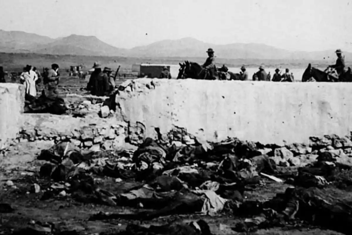 Fotografía en blanco y negro de un sitio devastado con cuerpos en el suelo, una pared derruida y varias personas y caballos al fondo en un paisaje árido con montañas. Fotografía en blanco y negro de un sitio devastado con cuerpos en el suelo, una pared derruida y varias personas y caballos al fondo en un paisaje árido con montañas.