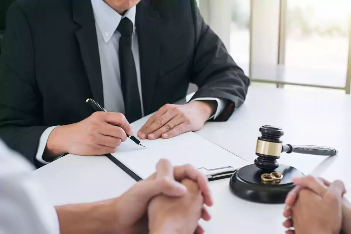 Tres personas sentadas en una mesa durante una reunión legal con un juez escribiendo en un documento y un mazo con dos anillos dorados sobre la mesa