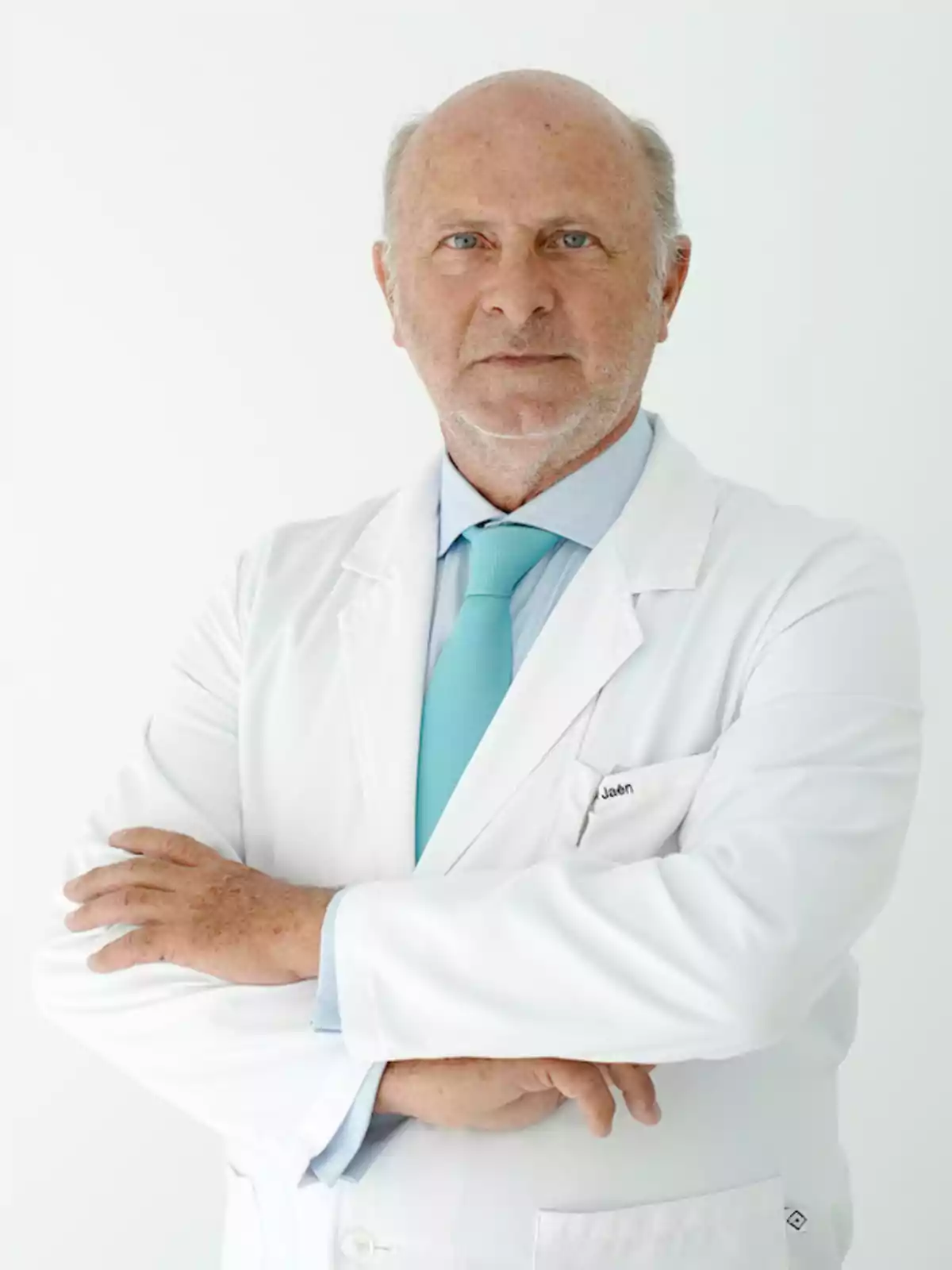 Hombre mayor con bata de médico y corbata celeste posando con los brazos cruzados sobre un fondo blanco Hombre mayor con bata de médico y corbata celeste posando con los brazos cruzados sobre un fondo blanco