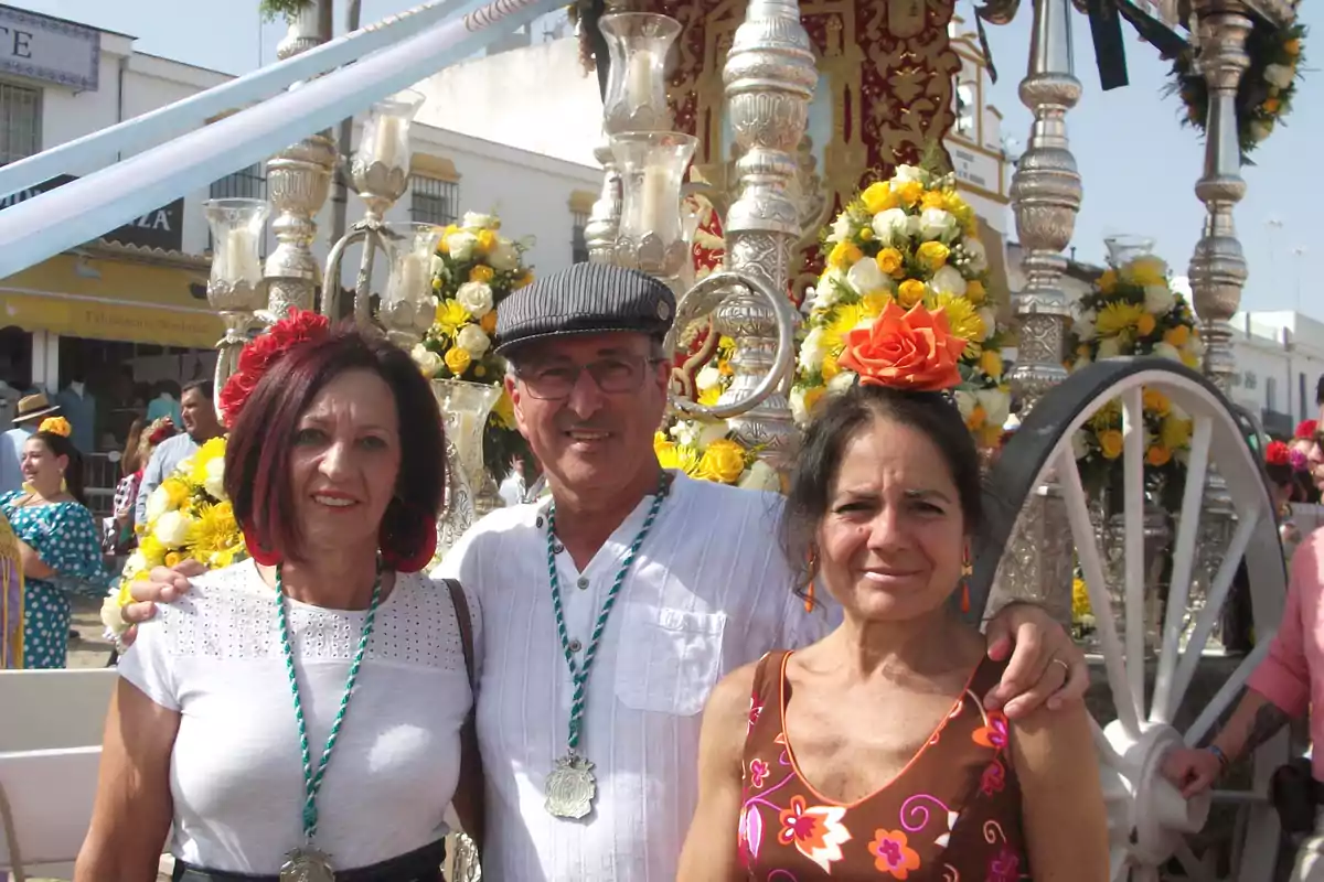 Tres personas sonríen vestidas con ropa tradicional andaluza frente a una carreta decorada con flores amarillas y detalles plateados durante una fiesta popular Tres personas sonríen vestidas con ropa tradicional andaluza frente a una carreta decorada con flores amarillas y detalles plateados durante una fiesta popular