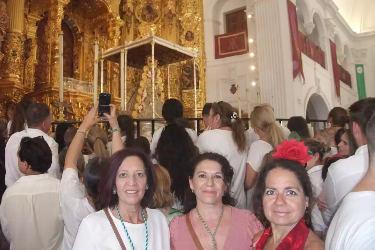 Tres mujeres posan sonrientes en primer plano durante una celebración religiosa en una iglesia decorada con detalles dorados y rodeadas de una multitud vestida de blanco Tres mujeres posan sonrientes en primer plano durante una celebración religiosa en una iglesia decorada con detalles dorados y rodeadas de una multitud vestida de blanco