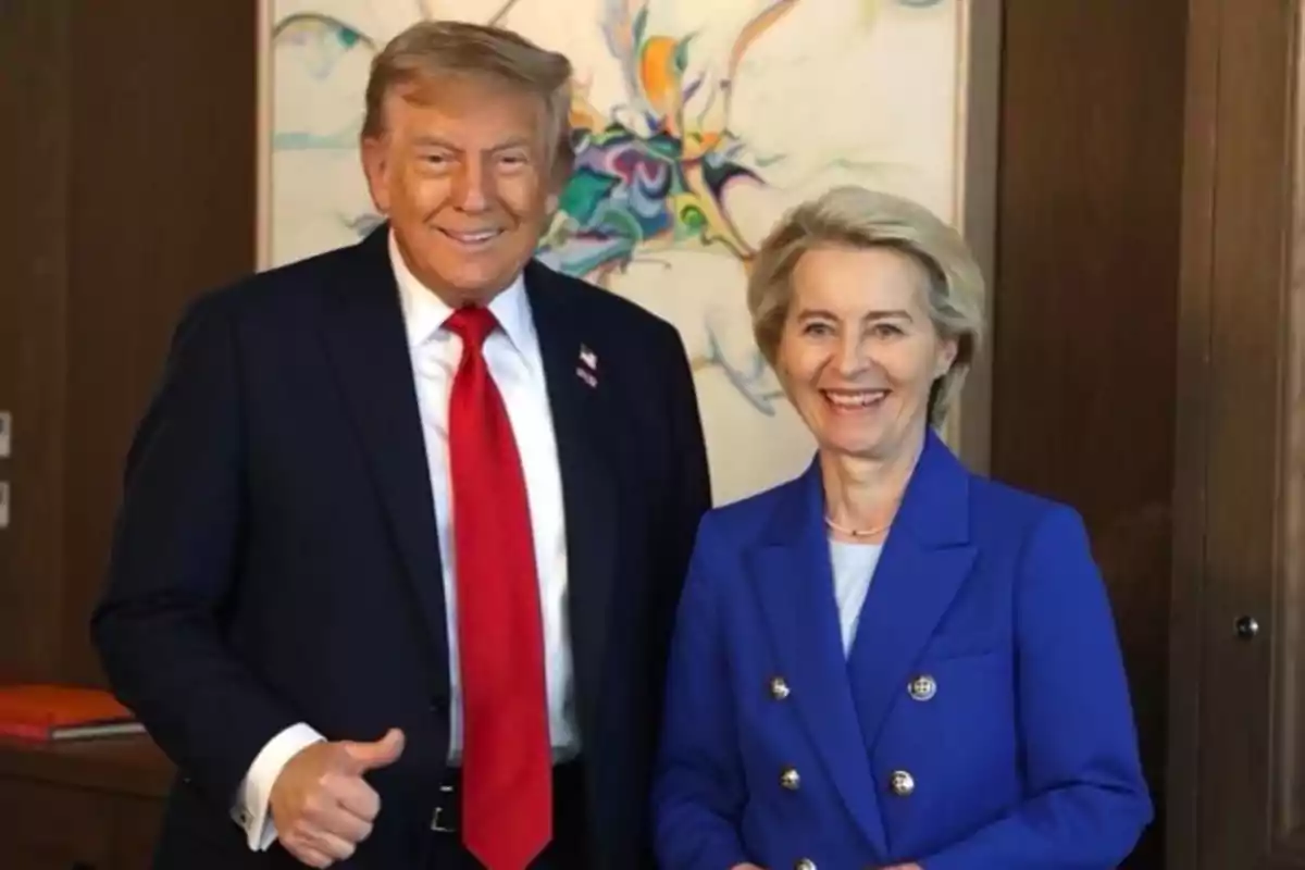 Dos personas de pie sonriendo, una de ellas lleva traje oscuro con corbata roja y la otra viste un saco azul, al fondo hay una pintura colorida abstracta. Dos personas de pie sonriendo, una de ellas lleva traje oscuro con corbata roja y la otra viste un saco azul, al fondo hay una pintura colorida abstracta.