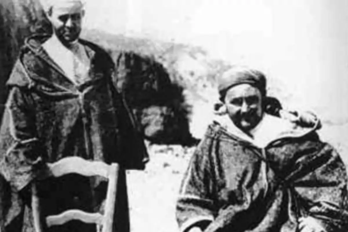 Dos personas con vestimenta tradicional posan al aire libre junto a una silla en una fotografía antigua en blanco y negro Dos personas con vestimenta tradicional posan al aire libre junto a una silla en una fotografía antigua en blanco y negro