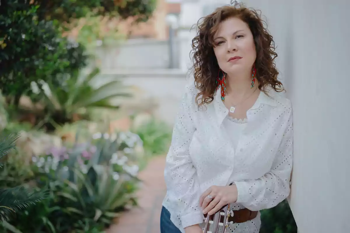 Mujer de cabello rizado y castaño con blusa blanca apoyada en una pared en un jardín Mujer de cabello rizado y castaño con blusa blanca apoyada en una pared en un jardín