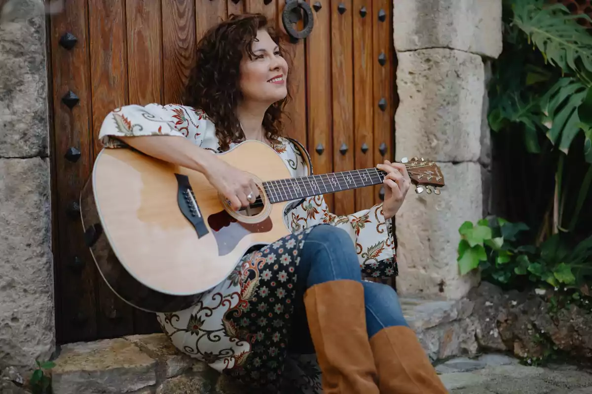 Mujer sentada tocando una guitarra acústica frente a una puerta de madera antigua con plantas a su alrededor