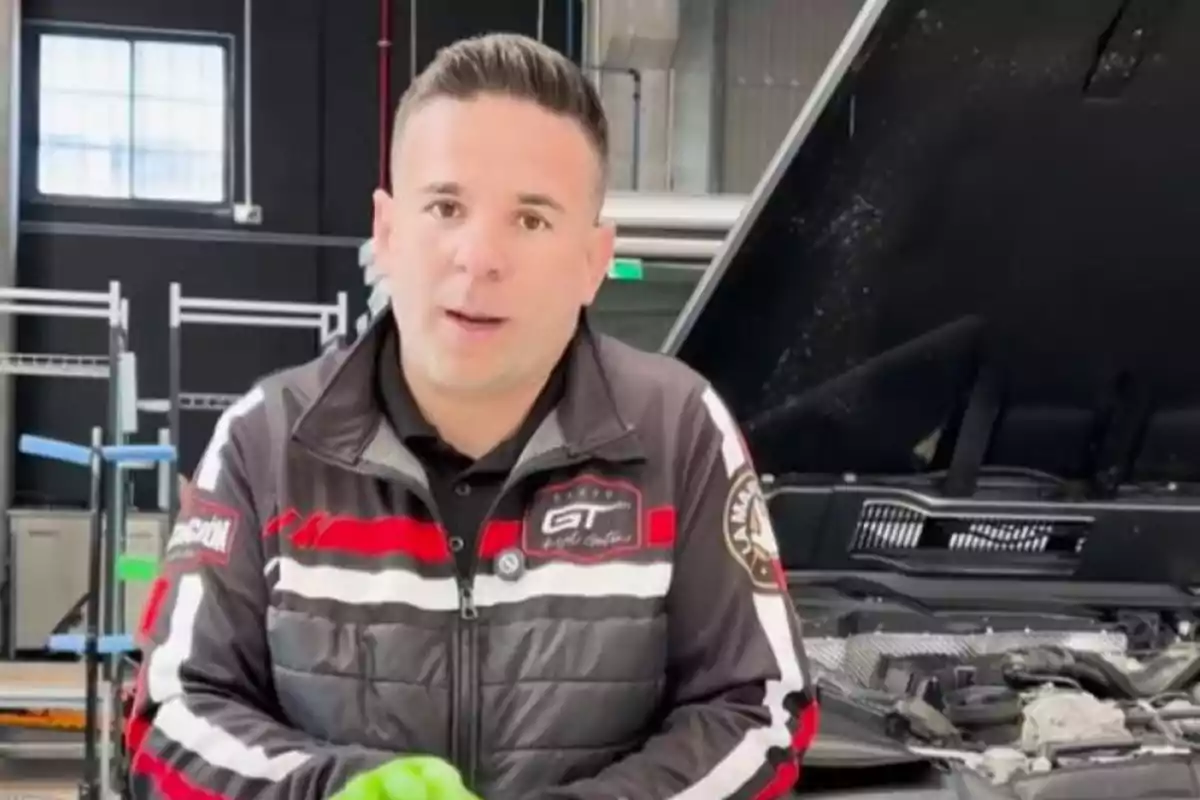 Hombre con chaqueta deportiva negra y roja trabajando en un taller mecánico frente a un auto con el cofre abierto