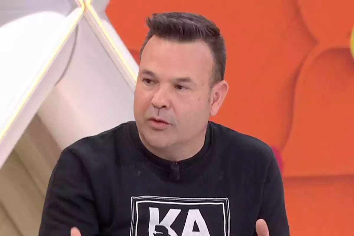Hombre de cabello corto y oscuro con camiseta negra participando en un programa de televisión con fondo naranja Hombre de cabello corto y oscuro con camiseta negra participando en un programa de televisión con fondo naranja