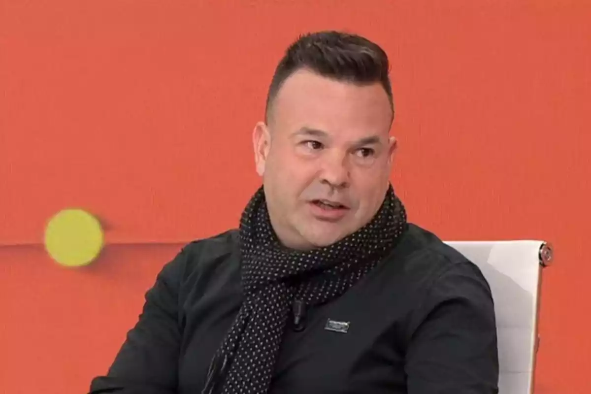 Hombre de cabello corto y oscuro con bufanda negra y camisa negra sentado en una silla blanca frente a un fondo naranja Hombre de cabello corto y oscuro con bufanda negra y camisa negra sentado en una silla blanca frente a un fondo naranja