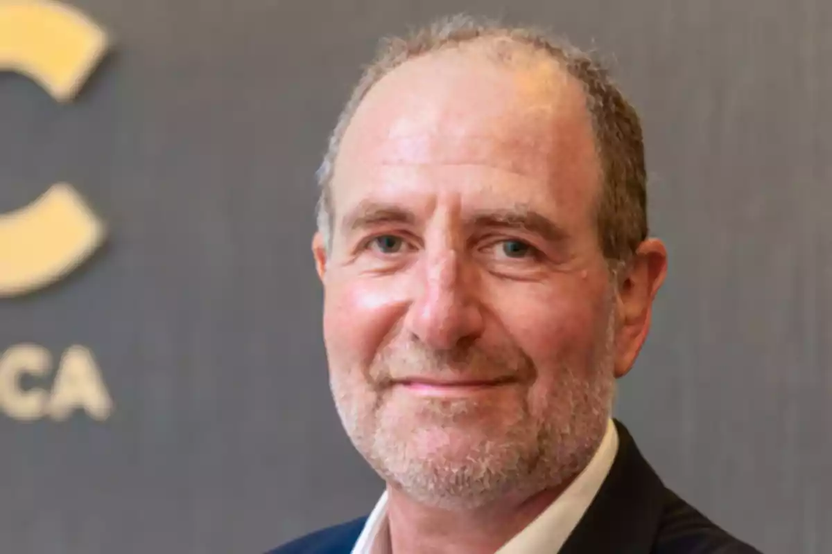 Hombre de mediana edad con barba y cabello corto, vestido con saco oscuro y camisa clara, sonriendo frente a un fondo gris con letras desenfocadas. Hombre de mediana edad con barba y cabello corto, vestido con saco oscuro y camisa clara, sonriendo frente a un fondo gris con letras desenfocadas.