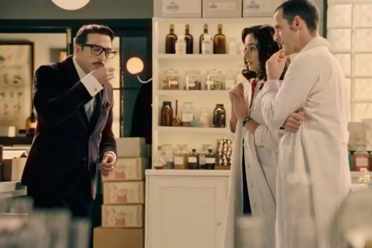 Tres personas en un laboratorio, dos con bata blanca observan a un hombre de traje -que es Bruno Tamarit- que prueba el contenido de una taza.