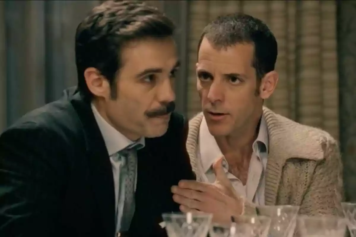 Dos hombres conversan en una mesa con copas de cristal al frente, uno de ellos lleva bigote -el cual es Guillermo Barrientos- y traje mientras el otro viste un suéter beige y parece estar hablando seriamente.