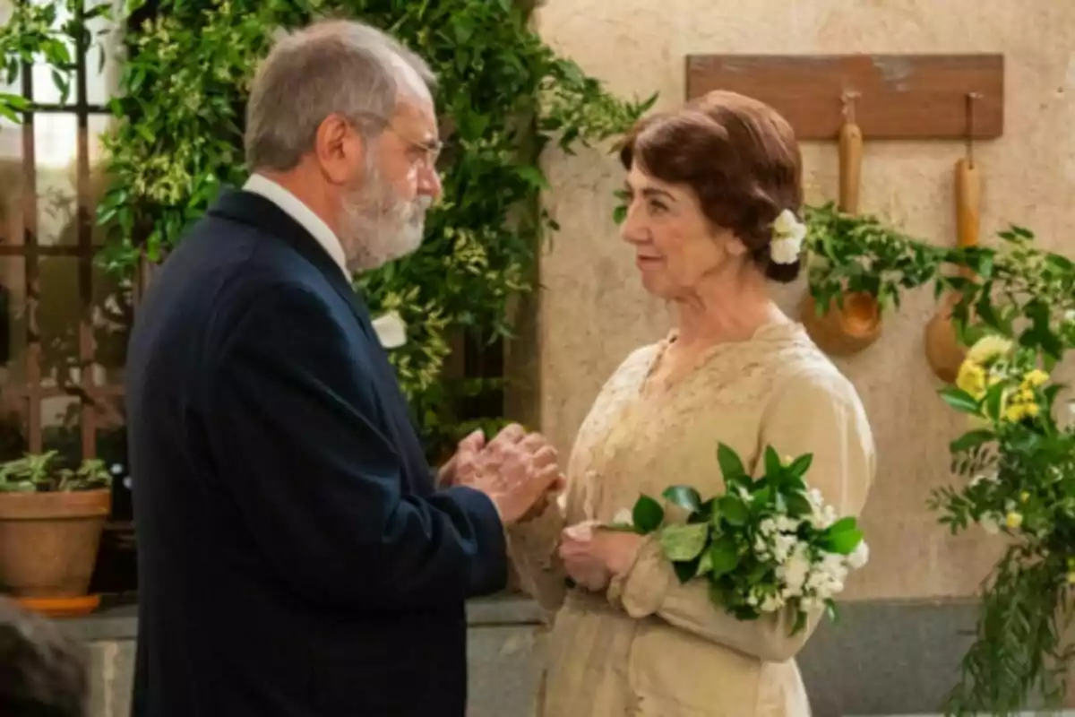 Pareja de personas mayores, que son Rómulo y Emilia en 'La Promesa', vestidas de manera elegante mirándose con cariño en un ambiente decorado con plantas y utensilios de cocina, la mujer sostiene un ramo de flores blancas y verdes. Pareja de personas mayores, que son Rómulo y Emilia en 'La Promesa', vestidas de manera elegante mirándose con cariño en un ambiente decorado con plantas y utensilios de cocina, la mujer sostiene un ramo de flores blancas y verdes.
