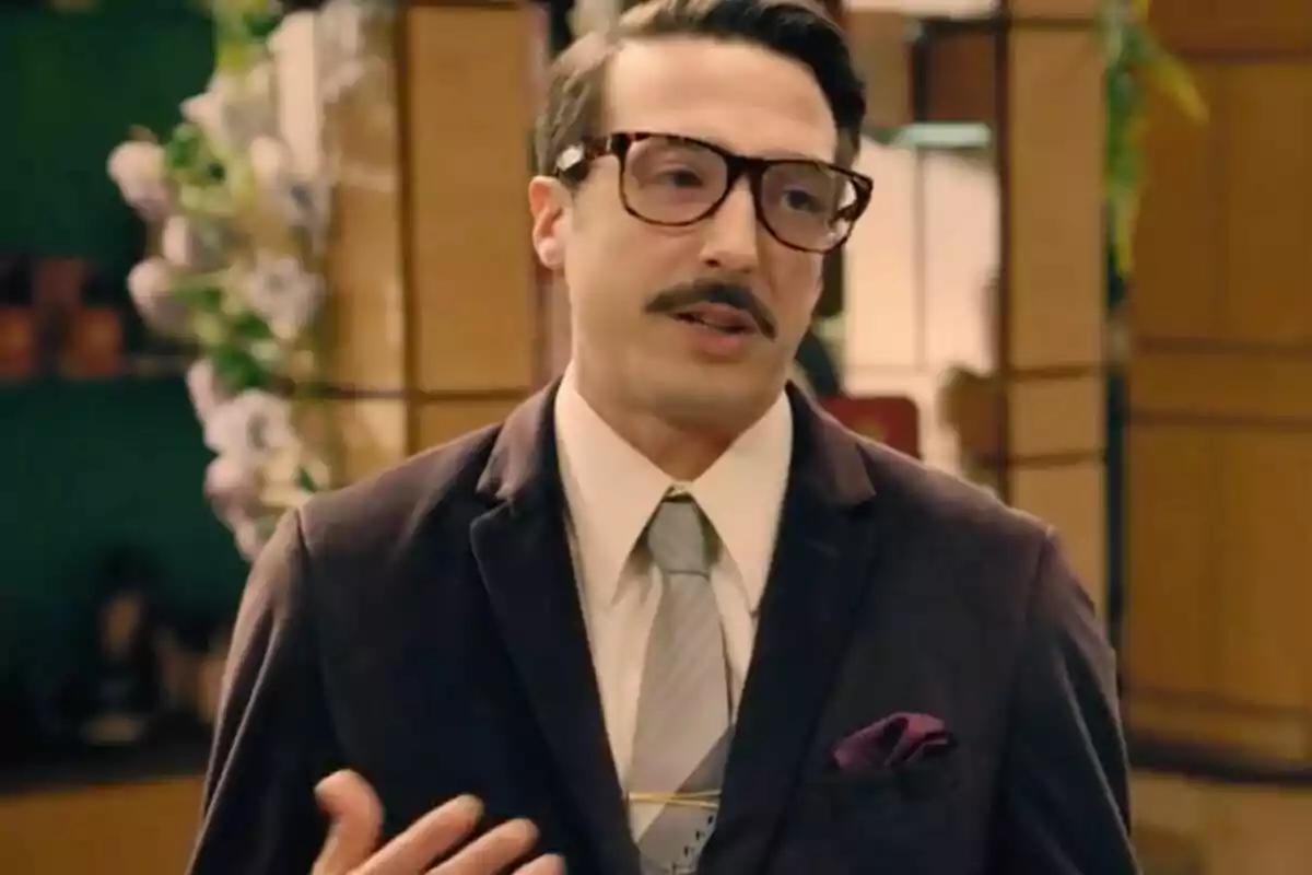 Cobeaga con gafas grandes y bigote vestido con traje oscuro y corbata clara en un ambiente interior elegante.