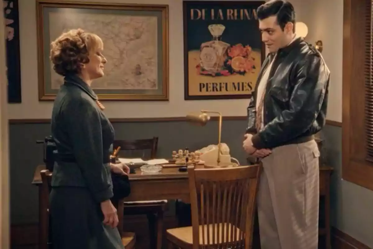 Una mujer y un hombre, que son Irene y Tasío, conversan en una oficina decorada con un mapa antiguo y un cartel de perfumes, ambos vestidos con ropa de época.