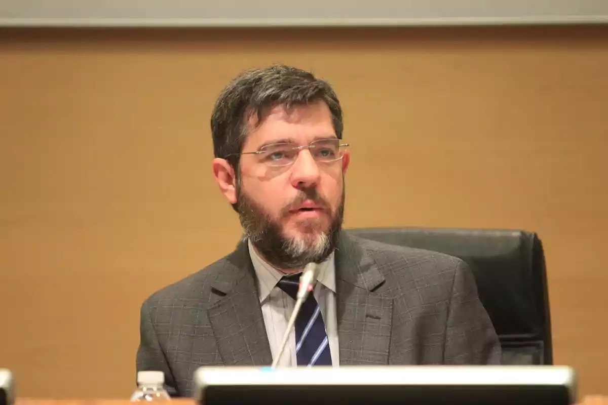 Hombre con barba y gafas hablando frente a un micrófono en una sala de conferencias Hombre con barba y gafas hablando frente a un micrófono en una sala de conferencias