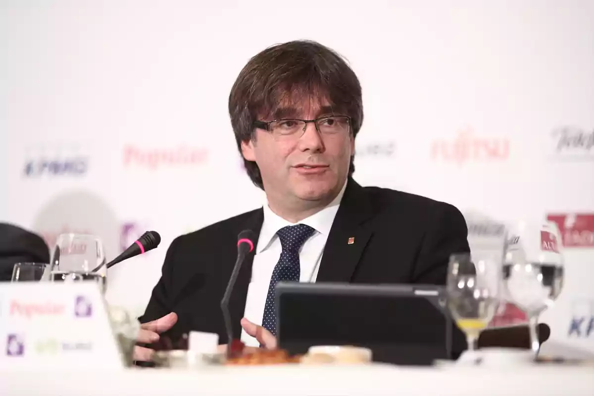 Hombre con gafas y traje oscuro hablando en una conferencia con micrófono y copas de cristal sobre la mesa Hombre con gafas y traje oscuro hablando en una conferencia con micrófono y copas de cristal sobre la mesa