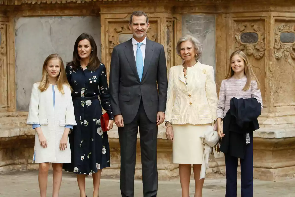 Cinco personas posan juntas frente a una pared de piedra antigua, todas vestidas de manera elegante y formal