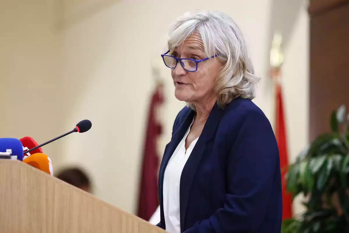 Mujer de cabello canoso y gafas azules hablando en un podio con micrófonos y fondo institucional Mujer de cabello canoso y gafas azules hablando en un podio con micrófonos y fondo institucional