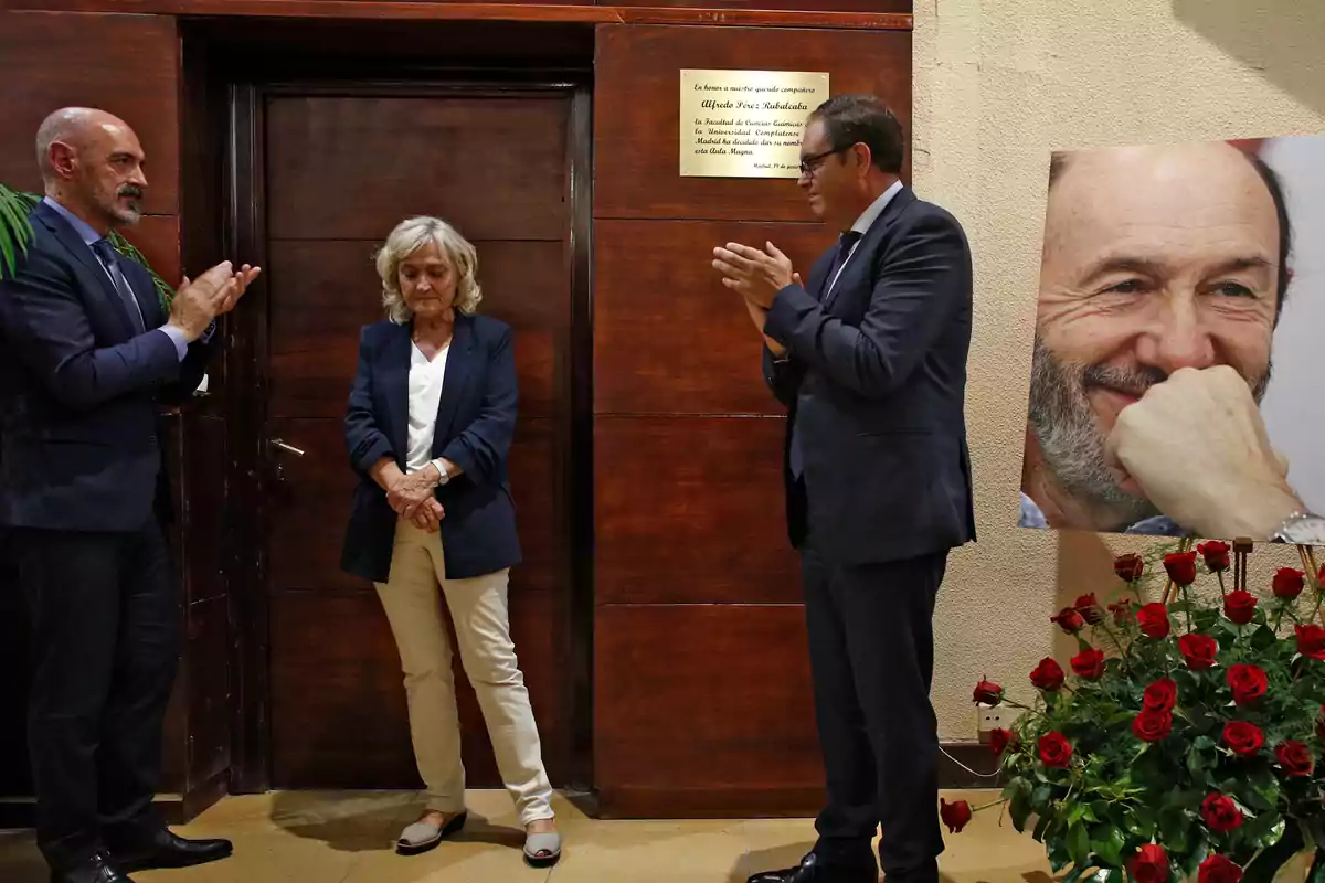 Tres personas vestidas formalmente participan en un homenaje junto a una placa conmemorativa y un retrato de un hombre sonriente rodeado de rosas rojas Tres personas vestidas formalmente participan en un homenaje junto a una placa conmemorativa y un retrato de un hombre sonriente rodeado de rosas rojas