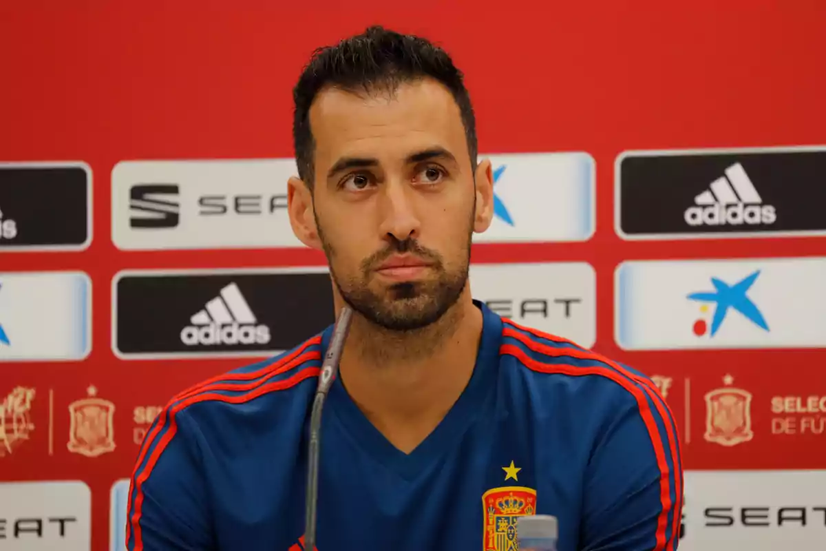 Futbolista con camiseta de la selección de España en una rueda de prensa frente a un fondo con logotipos de patrocinadores Futbolista con camiseta de la selección de España en una rueda de prensa frente a un fondo con logotipos de patrocinadores