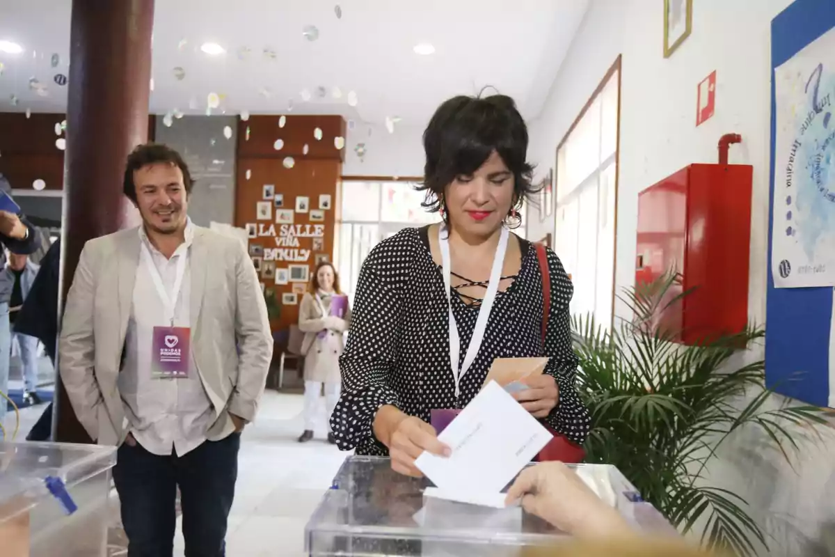 Mujer depositando su voto en una urna transparente mientras un hombre la observa y otras personas esperan en el fondo en un centro de votación Mujer depositando su voto en una urna transparente mientras un hombre la observa y otras personas esperan en el fondo en un centro de votación