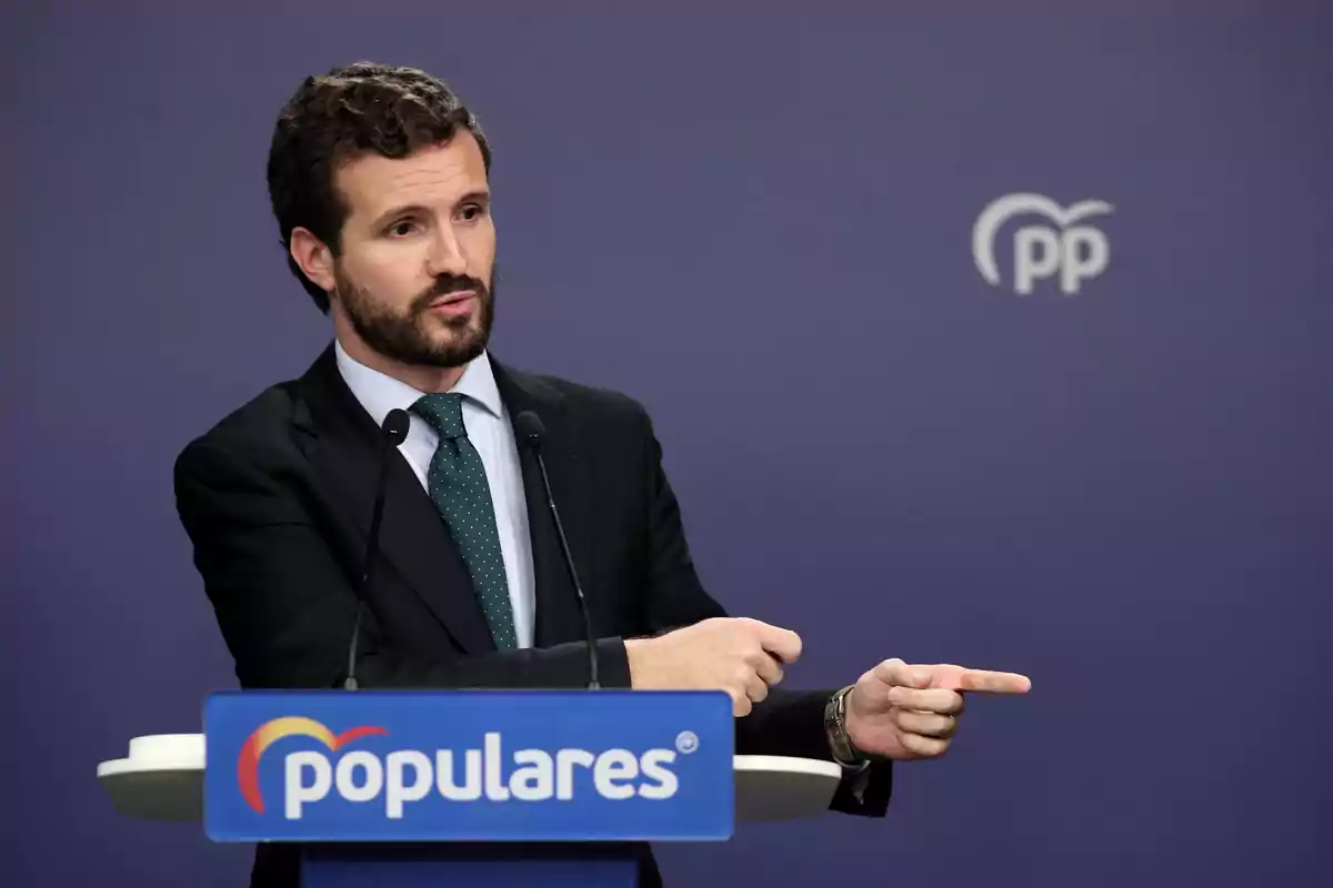 Hombre con barba y traje oscuro hablando en una rueda de prensa frente a un atril azul con el logo y la palabra populares sobre un fondo azul