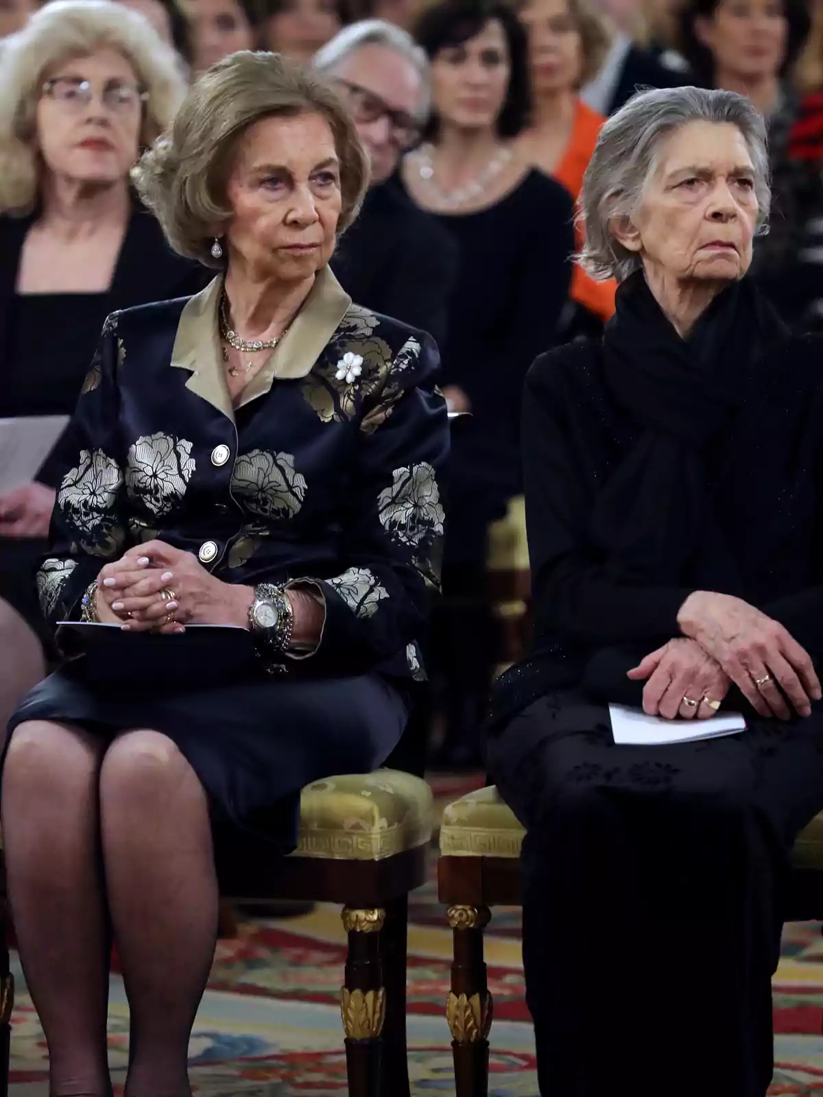 Dos mujeres mayores elegantemente vestidas están sentadas en primera fila durante un evento formal, rodeadas de otras personas en el público. Dos mujeres mayores elegantemente vestidas están sentadas en primera fila durante un evento formal, rodeadas de otras personas en el público.