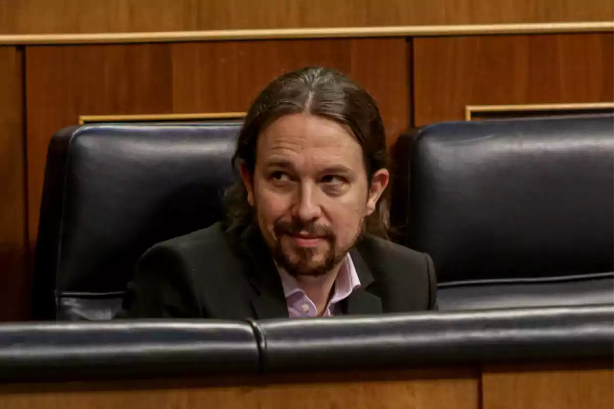 Hombre de cabello largo y barba sentado en un escaño de un parlamento con fondo de madera Hombre de cabello largo y barba sentado en un escaño de un parlamento con fondo de madera