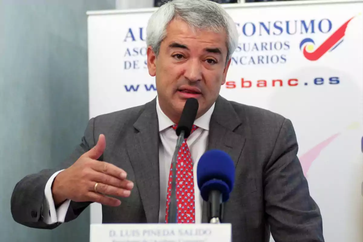 Hombre de cabello canoso con traje gris y corbata roja hablando frente a un micrófono en una conferencia de prensa Hombre de cabello canoso con traje gris y corbata roja hablando frente a un micrófono en una conferencia de prensa