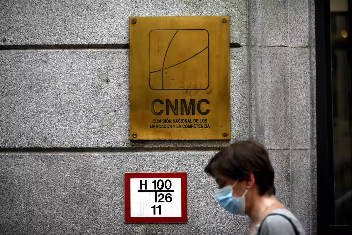 Placa dorada de la CNMC en una pared gris con una persona usando mascarilla caminando frente a ella Placa dorada de la CNMC en una pared gris con una persona usando mascarilla caminando frente a ella