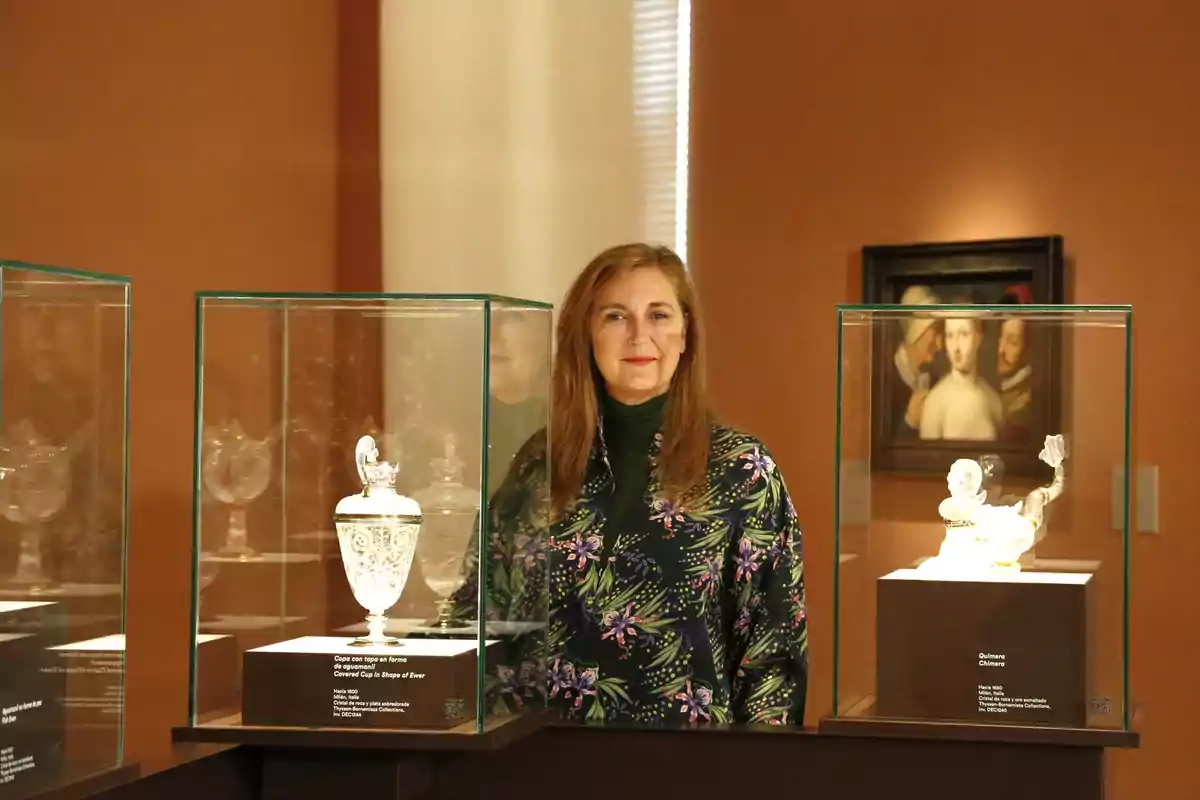 Mujer de cabello largo y castaño posando en una sala de museo entre vitrinas de cristal que exhiben piezas de arte decorativo, con paredes color terracota y un cuadro al fondo.