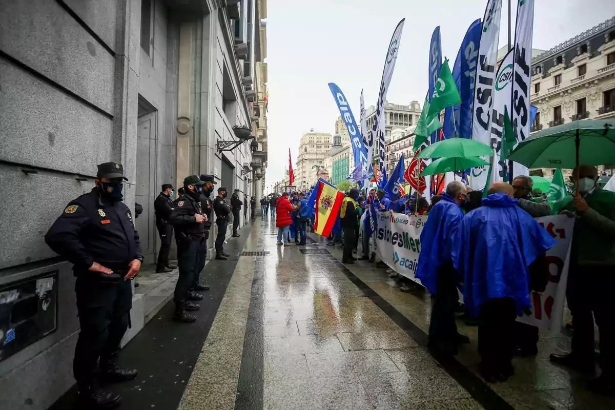Manifestación en la calle con personas portando pancartas y banderas bajo la lluvia mientras varios policías vigilan la escena desde la acera opuesta. Manifestación en la calle con personas portando pancartas y banderas bajo la lluvia mientras varios policías vigilan la escena desde la acera opuesta.