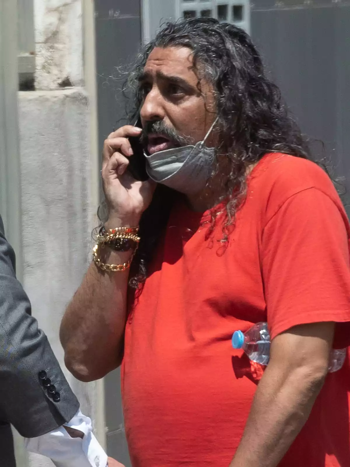Hombre de cabello largo y rizado con camiseta roja hablando por teléfono móvil y usando una mascarilla en la barbilla mientras sostiene una botella de agua bajo el brazo Hombre de cabello largo y rizado con camiseta roja hablando por teléfono móvil y usando una mascarilla en la barbilla mientras sostiene una botella de agua bajo el brazo