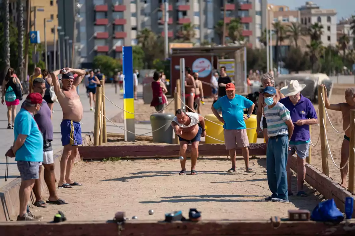 Un grupo de personas mayores juega a la petanca en una cancha de arena junto a la playa en un día soleado, mientras otras personas pasean por el paseo marítimo al fondo.